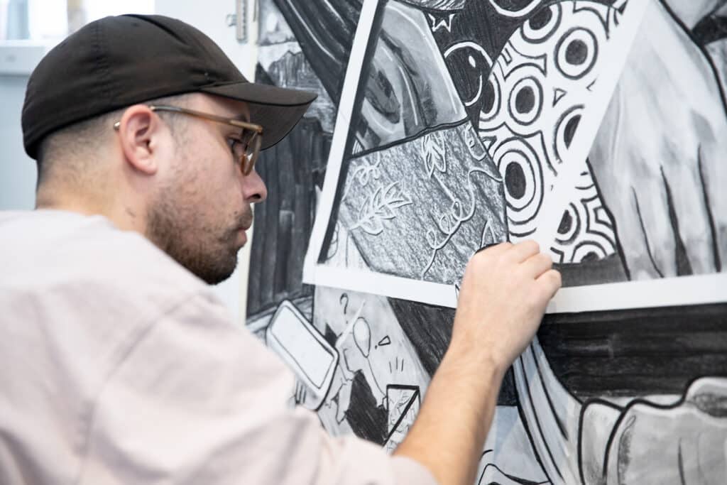 Close-up of a student wearing a cap concentrating while sketching details with charcoal on a large drawing.