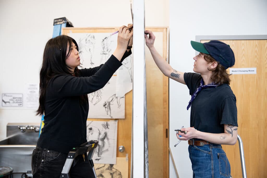 Two students working together on opposite sides of a drawing panel, one standing on a ladder and one on the floor.