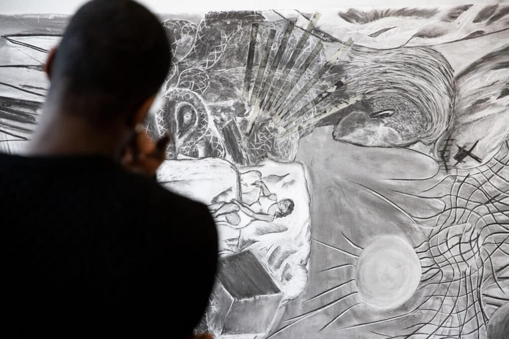 Close-up of a person’s back as they draw on a large charcoal mural featuring abstract shapes, textures, and a reclining figure.