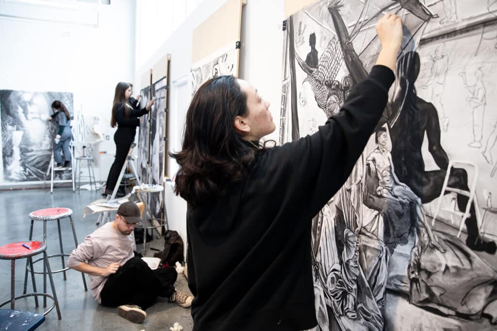Several students drawing large-scale works in a studio, some standing on ladders while others sit or crouch on the floor.
