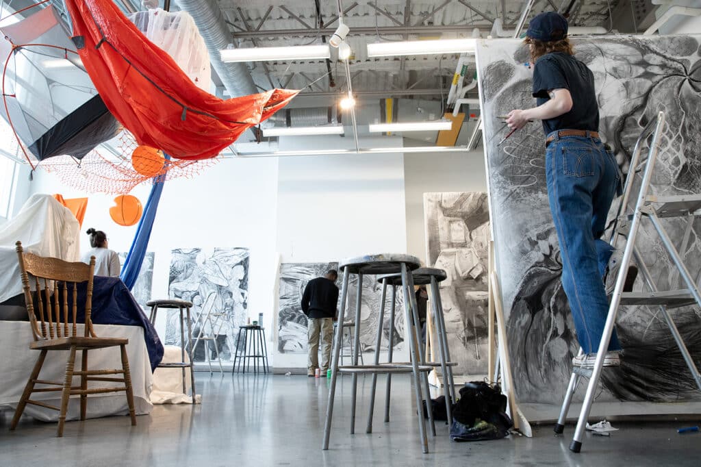 Wide studio view with fabric installations hanging overhead and students working on large-scale charcoal drawings along the walls.