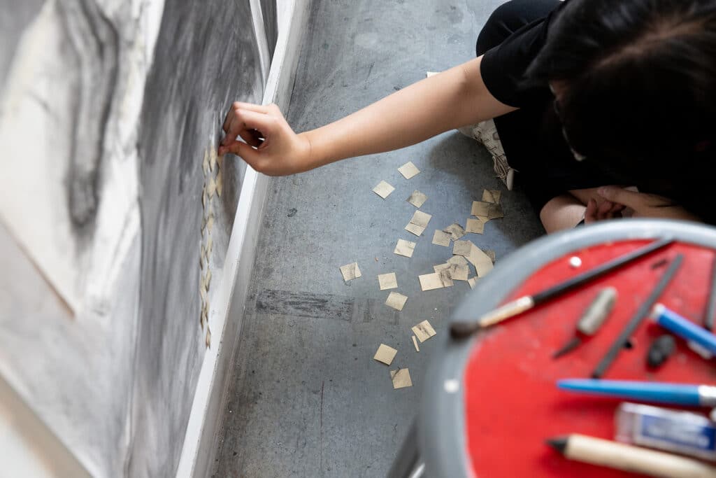 Close-up of a student’s hand pressing small paper squares onto a drawing near the floor, surrounded by scattered cut pieces.