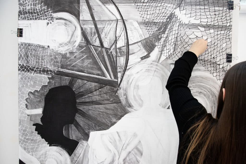 Close-up of a student’s hand drawing a net-like pattern with charcoal on a large black-and-white piece featuring silhouettes and abstract shapes.