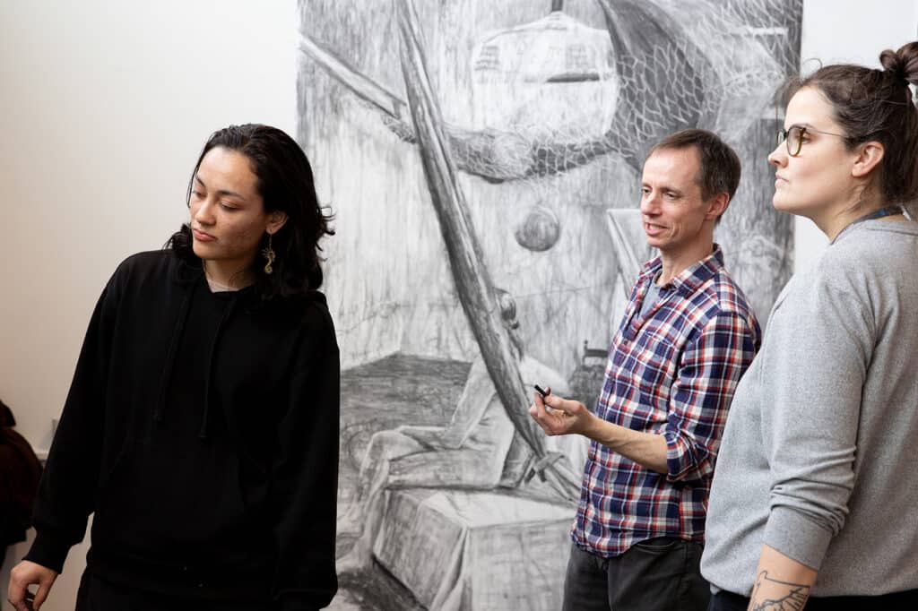 Three people stand in front of a large charcoal drawing during a critique session, with one person gesturing while holding a drawing tool.