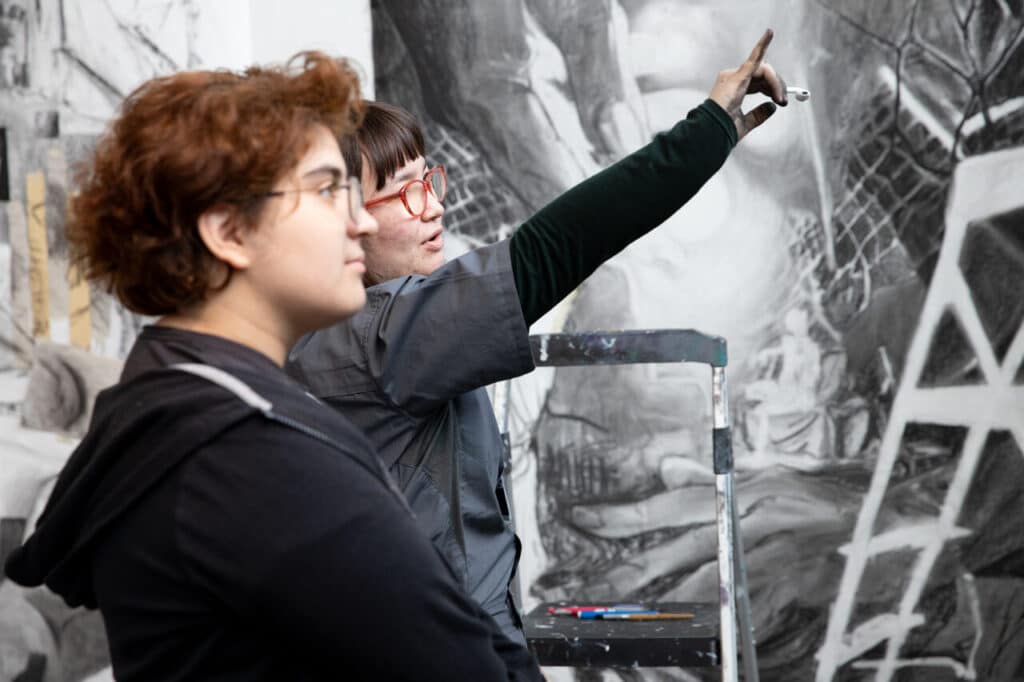 Two students in front of a large charcoal drawing; one gestures and points while explaining, the other looks on attentively.