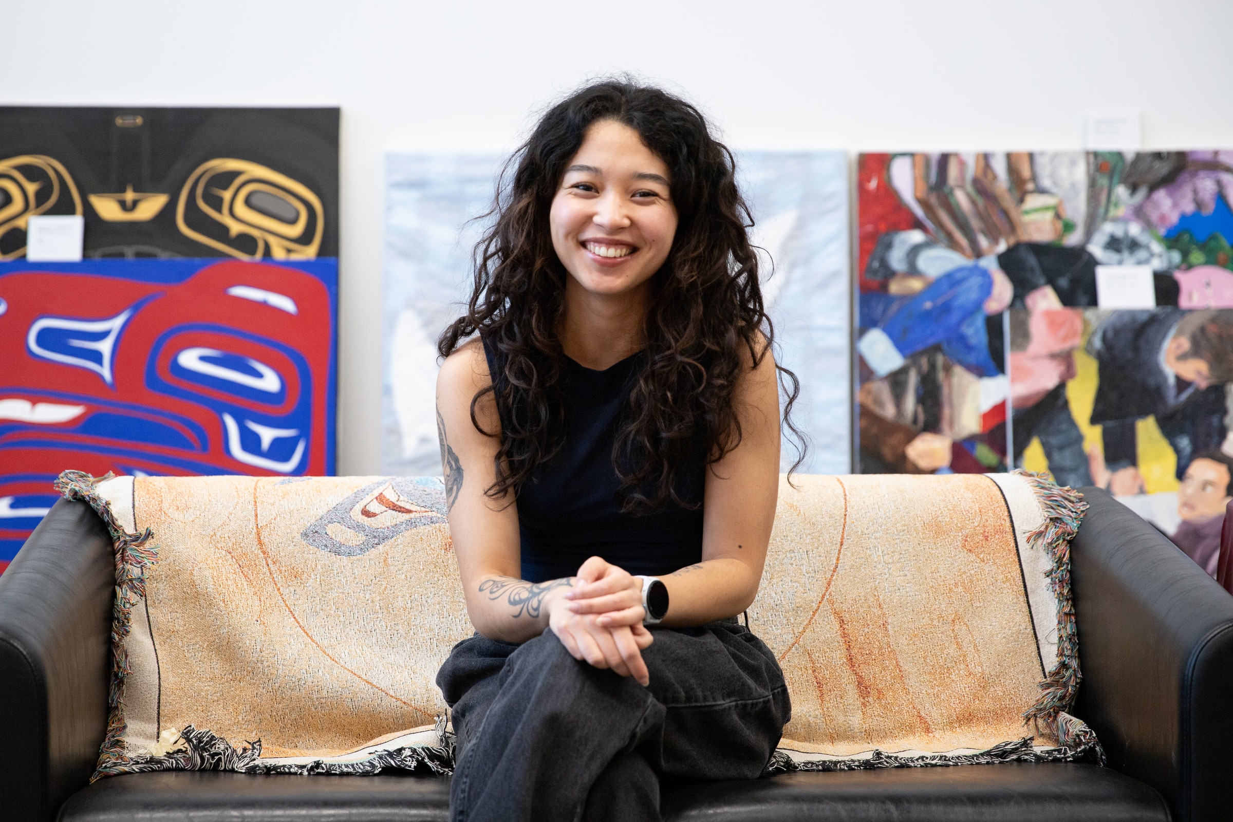 Visual artist Ella White sits smiling on a couch with student artwork displayed on the wall behind her at Emily Carr University.