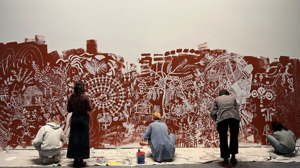 A group of people painting a large mural with white designs on a red background, featuring plants, animals and abstract patterns.
