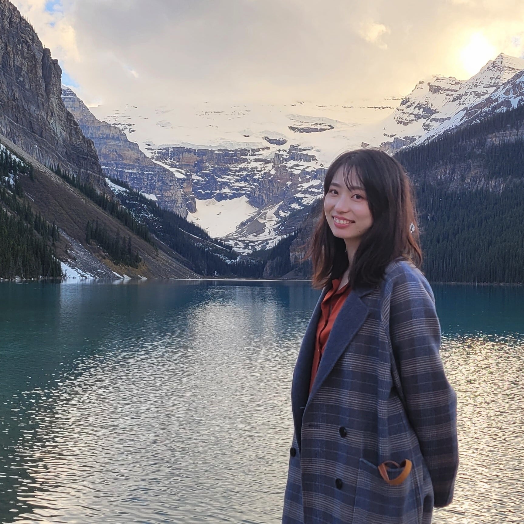 A person smiling while standing outdoors near a turquoise mountain lake, with snow-covered peaks and forested slopes in the background at sunset.