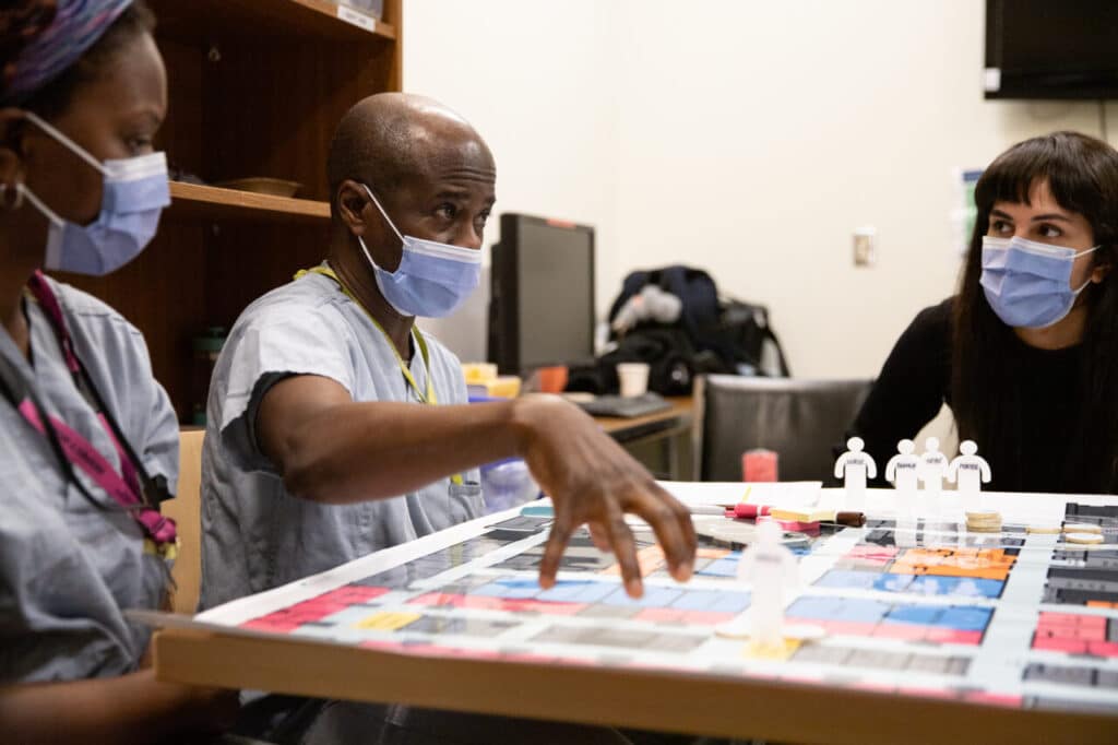 People in scrubs and masks discussing around a board with paper figures labelled “NURSE,” “PORTER,” and “SURRAY.”