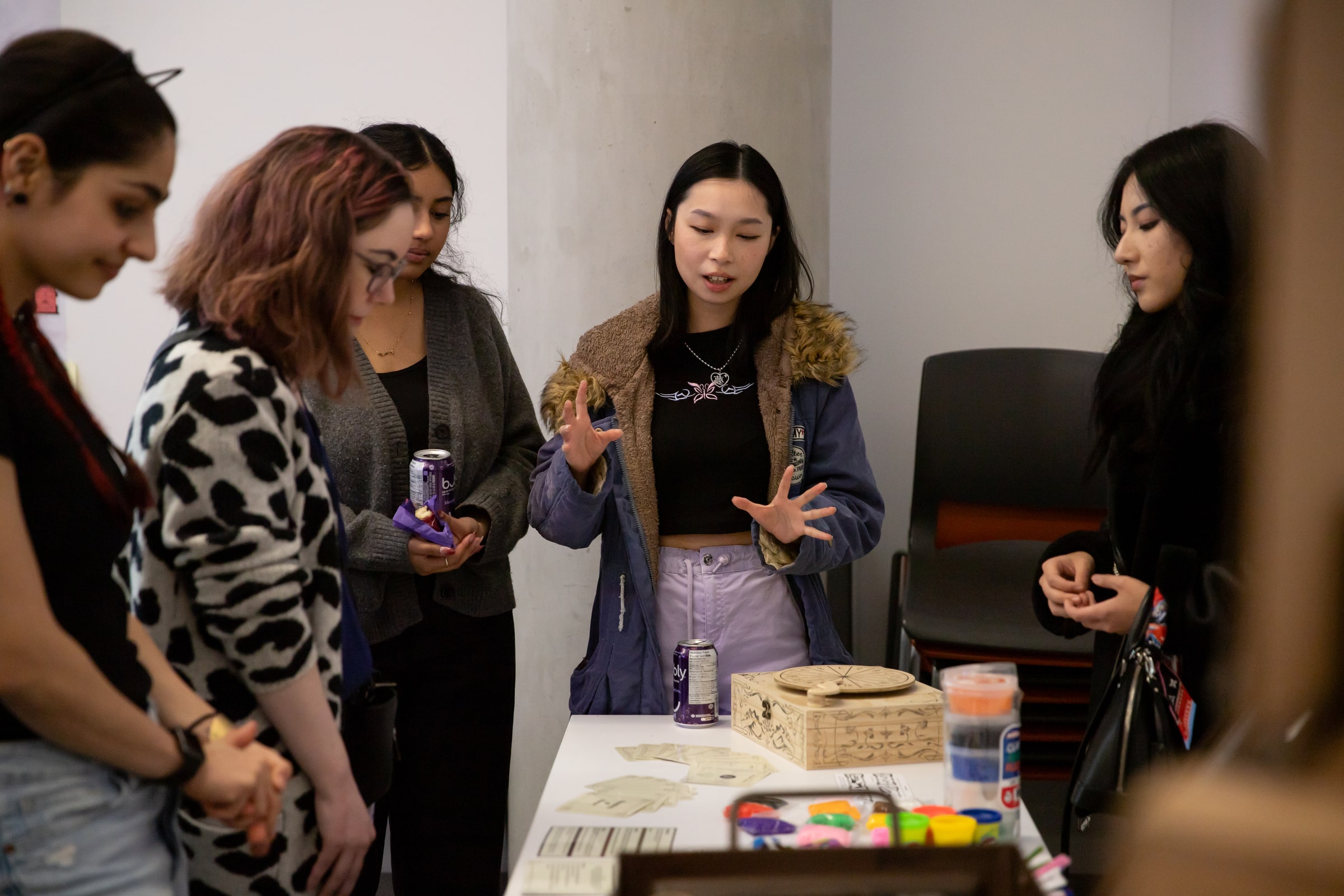 A student presents to a small group, standing at a table with illustrated materials and colorful supplies while others listen.