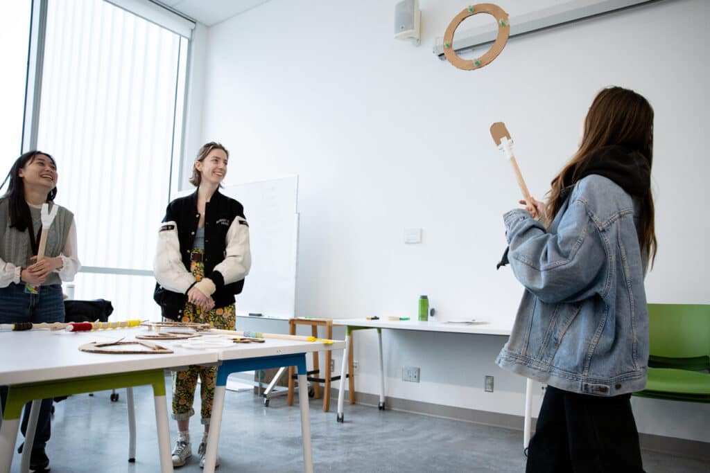 A student smiles while holding a stick topped with a large cardboard disc decorated with long, colourful ribbons.