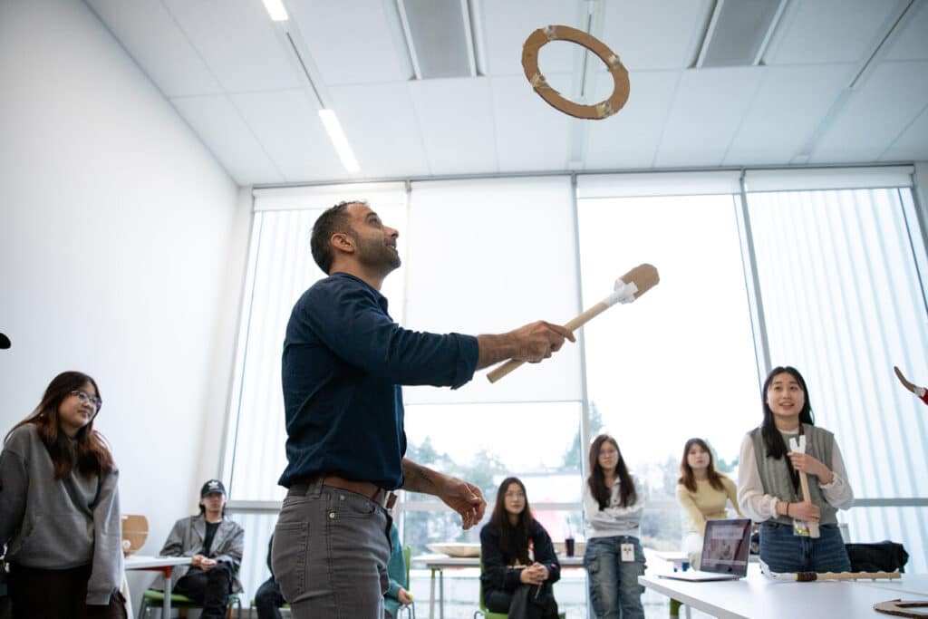 A person uses a stick with a cardboard attachment to toss a ring through a cardboard hoop, while students watch and smile.