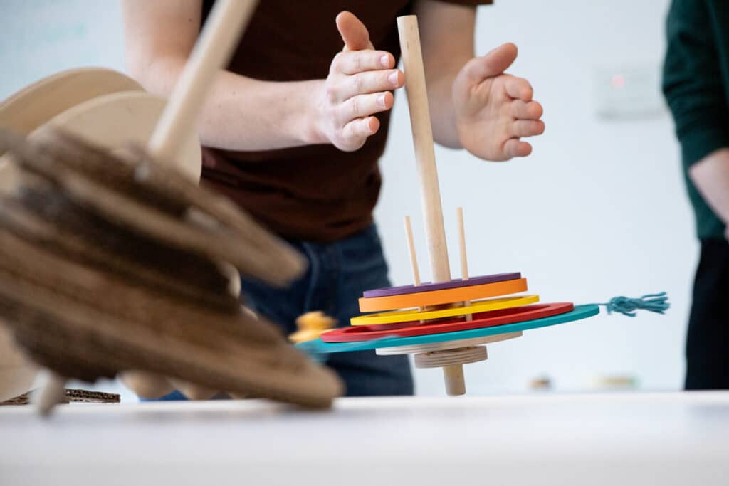 Close-up of colourful circular wooden pieces stacked on a vertical dowel, with hands reaching toward the object.