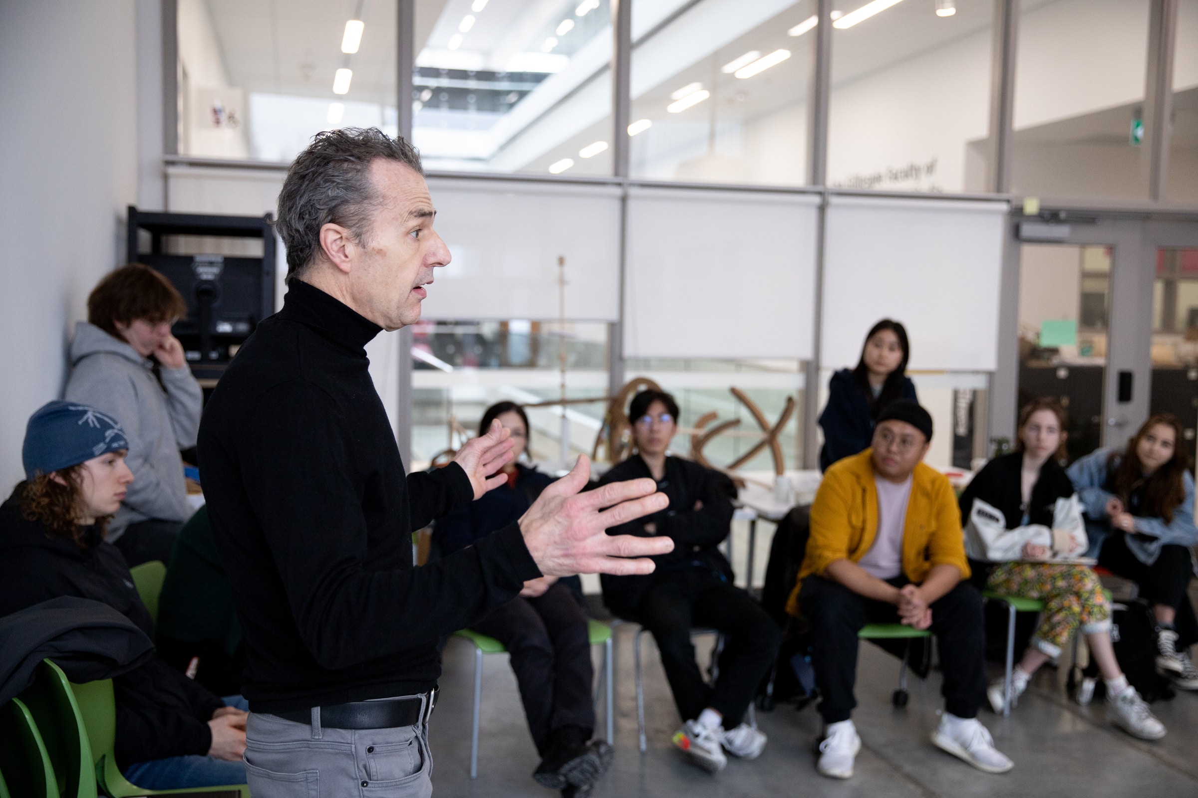 An instructor in a black turtleneck gestures with his hands while speaking to a group of students seated in a classroom. The students sit in green chairs, listening attentively, with some leaning forward and others watching from the back.