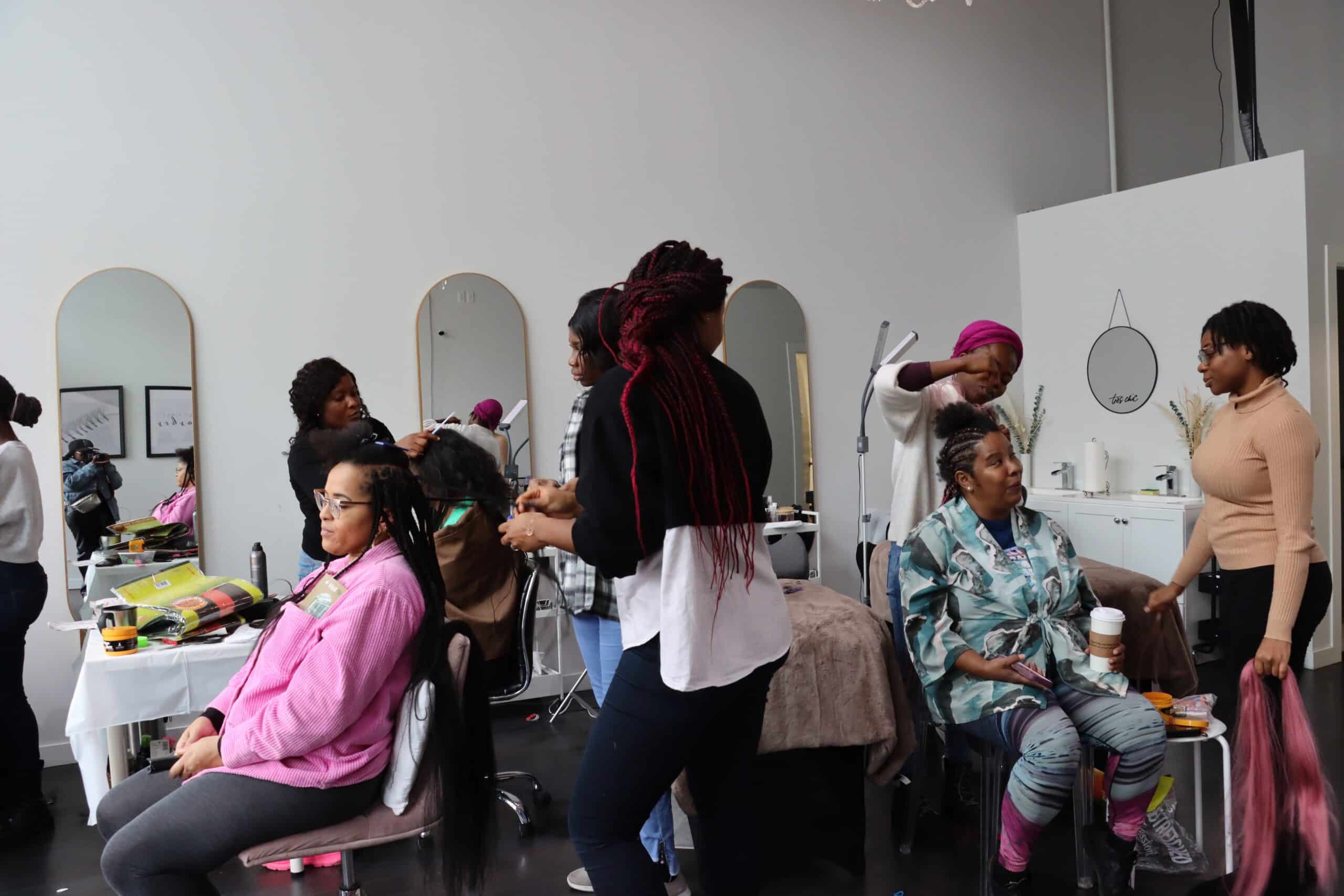 Busy hair salon scene with multiple women braiding and styling hair, while clients sit in chairs chatting and holding drinks.