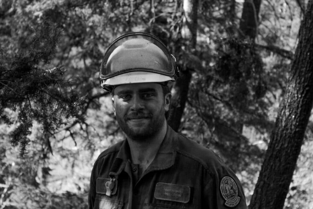 Smiling firefighter in protective gear and helmet, standing in the forest.