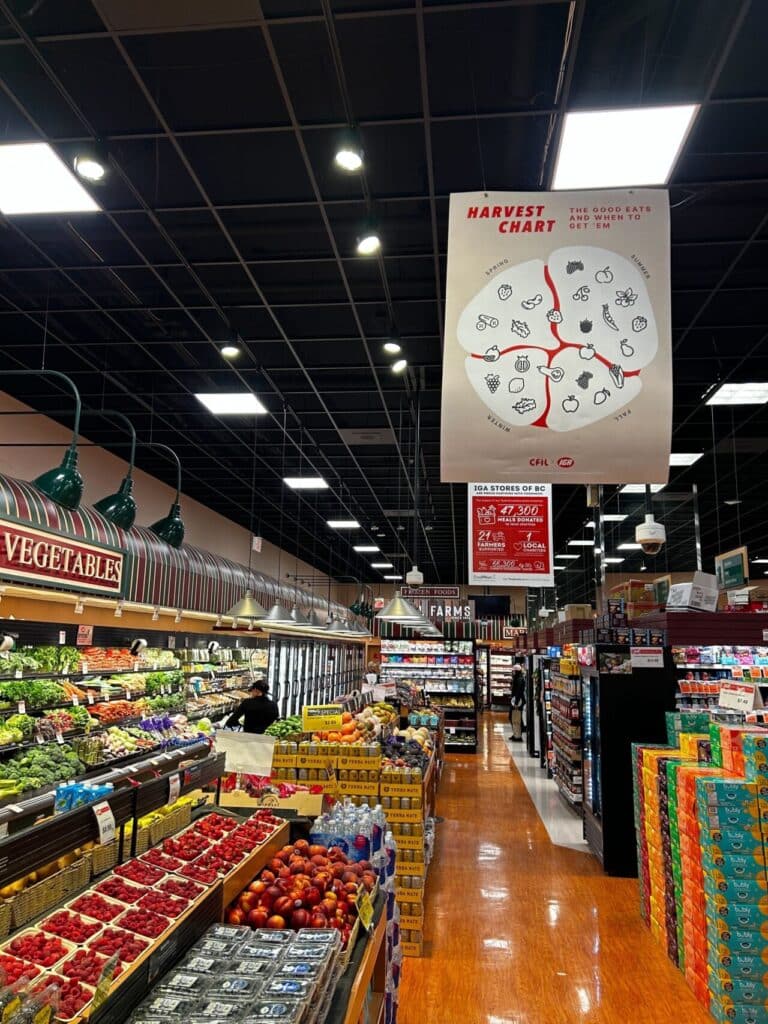 A grocery store produce aisle filled with fruits and vegetables, with a hanging sign overhead that reads “Harvest Chart: The Good Eats and When to Get ’Em.”
