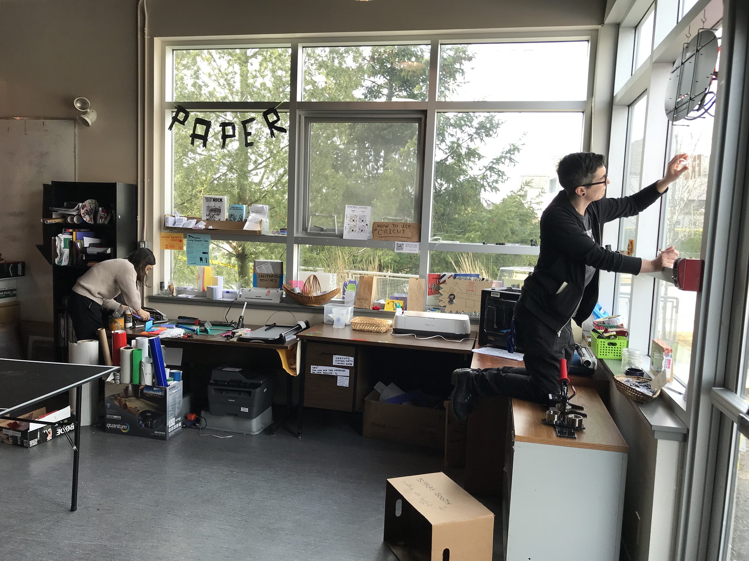 wo people working in a bright makerspace with large windows; one adjusts something on the window while the other cuts paper at a cluttered workstation labeled “PAPER.”