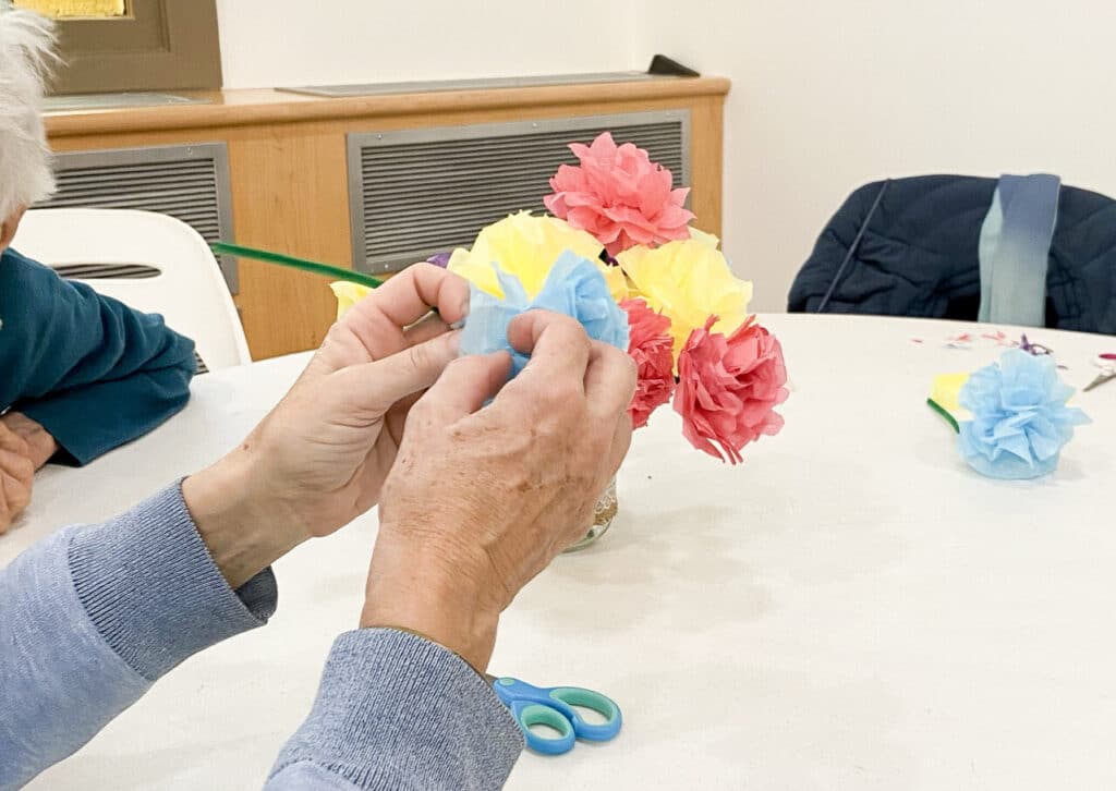 Hands crafting a blue tissue paper flower at a table, with a vase of completed red, yellow and pink paper flowers in the background.