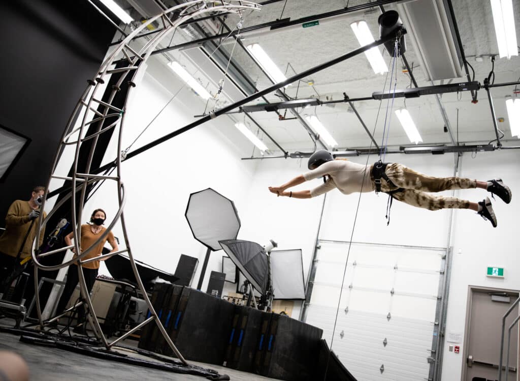 A person suspended in midair on a harness rig inside a studio, wearing a helmet and stretching forward as if flying, while two masked technicians observe from the ground.