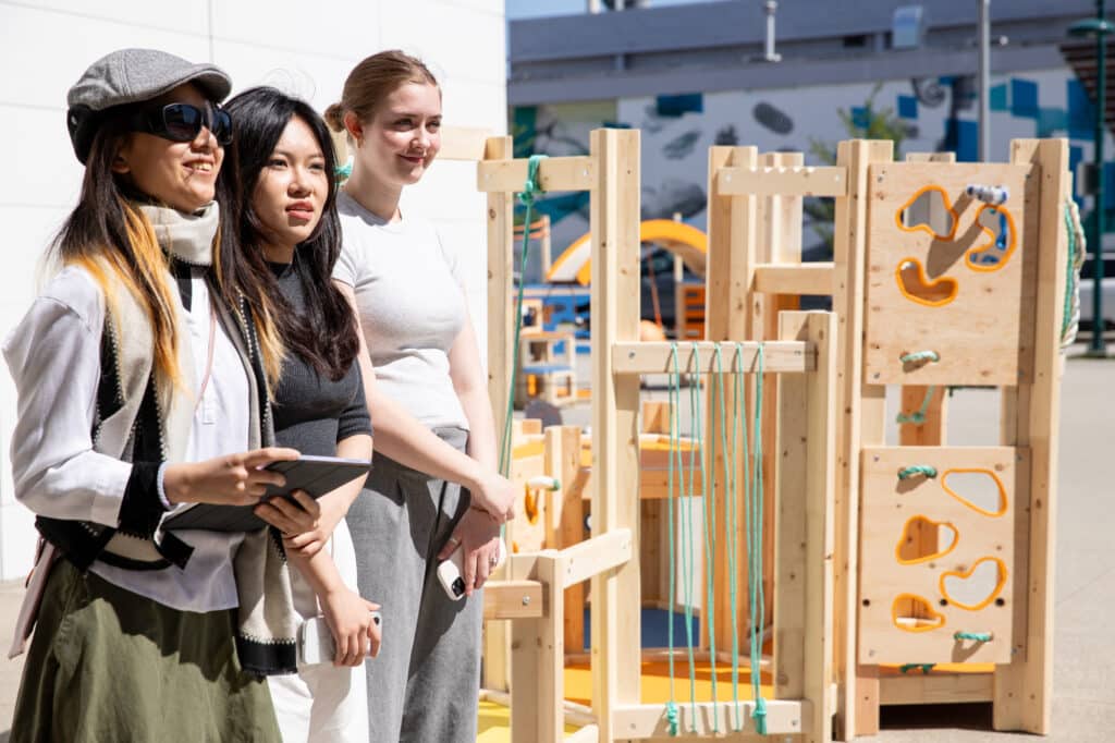 Three students stand outside beside a wooden playscape structure, smiling during their design presentation.