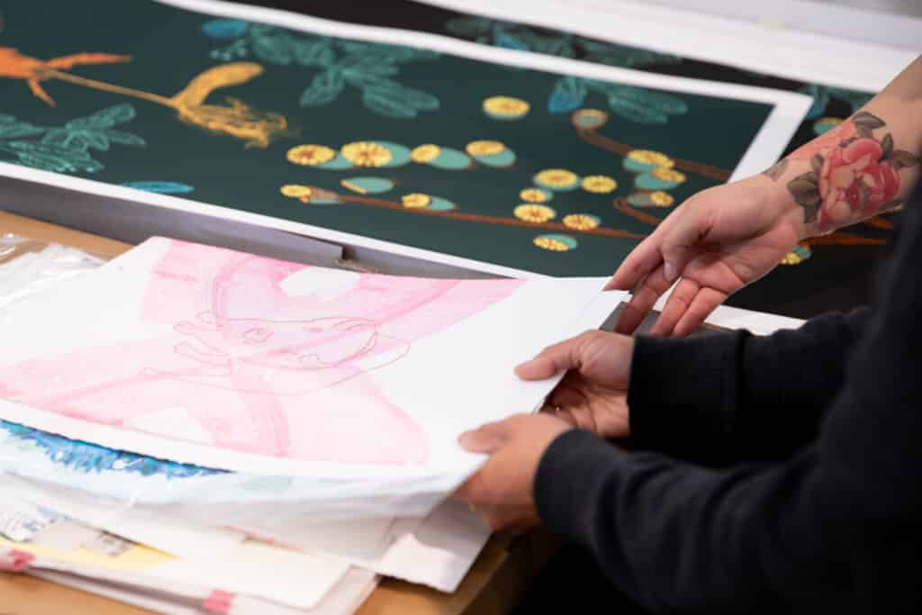 Hands holding a stack of colorful prints, with the top sheet showing a pale pink sketch and a large floral tattoo visible on one arm. A bold green print with yellow flowers lies on the table in the background.