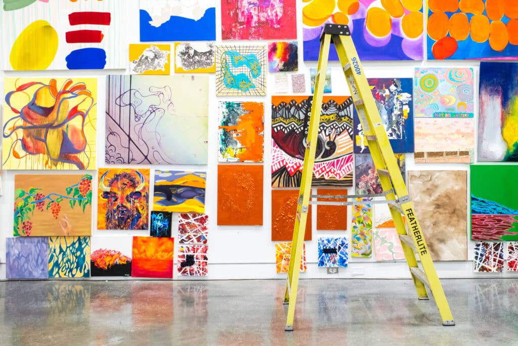 Brightly coloured paintings and mixed media works displayed on a wall, with a yellow ladder in front.