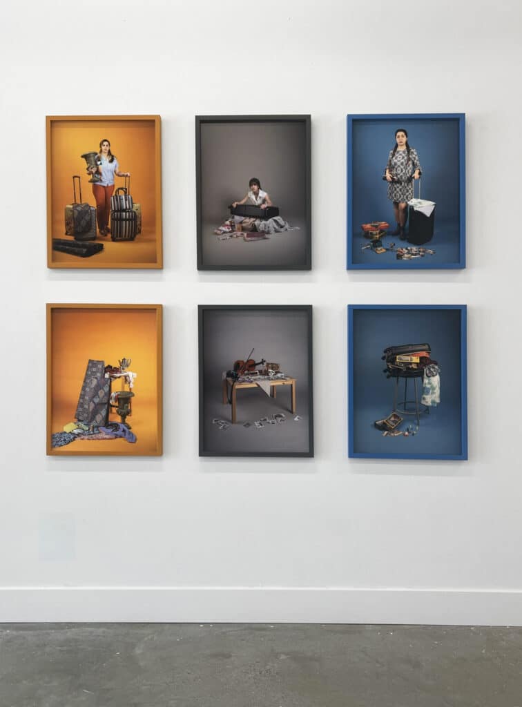 Six framed photographs hung in two rows on a gallery wall, each showing people with luggage or personal belongings arranged against brightly coloured backdrops.
