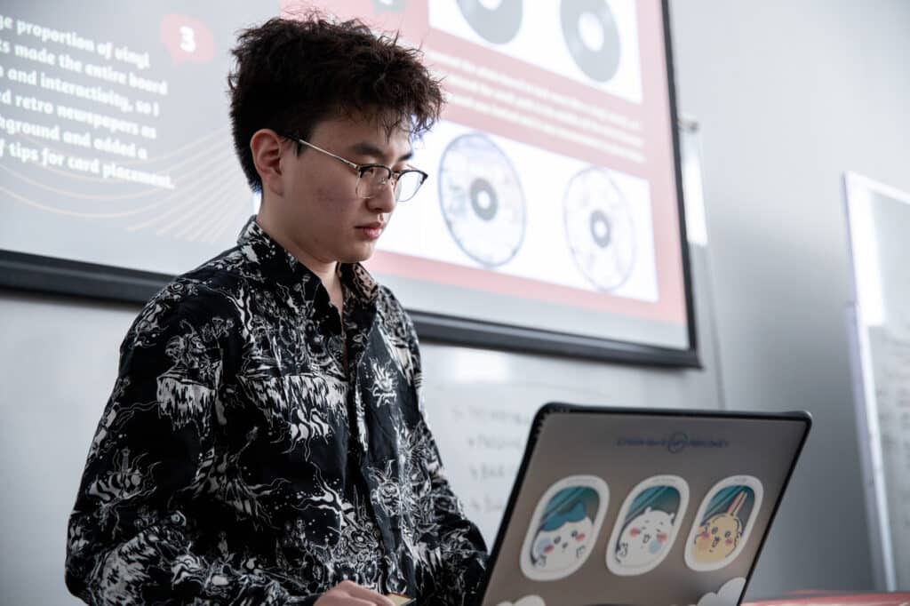 A student wearing glasses works on a laptop covered in cartoon stickers while giving a presentation. A large projected slide with images and text is visible behind him.