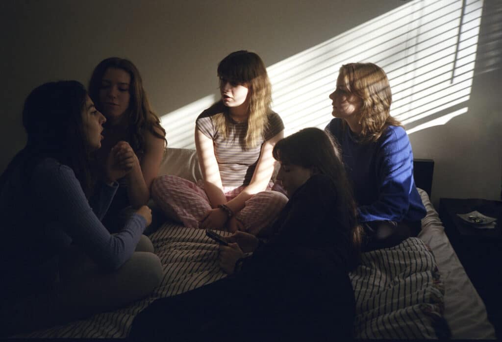 Five young people gathered on a bed in a dimly lit room, some talking while others sit quietly, with sunlight streaming through blinds casting striped shadows on the wall