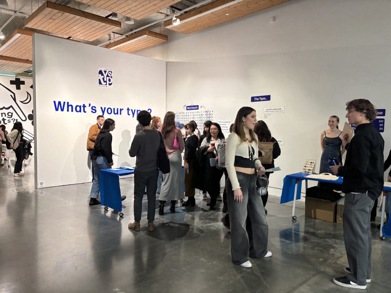 Visitors chat and browse interactive displays at a student-run typography exhibition with a large wall text reading “What’s your type?”
