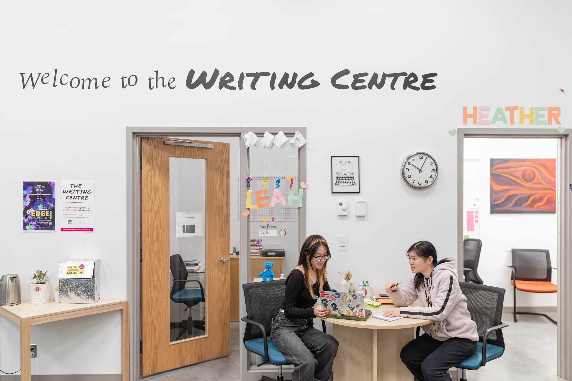 A bright interior of the Writing Centre at Emily Carr University. A large sign reads “Welcome to the Writing Centre” above a doorway. Two students sit at a round table in the foreground, one with a laptop covered in stickers, the other writing in a notebook. The walls are decorated with colorful paper name signs, a clock, posters, and artwork.