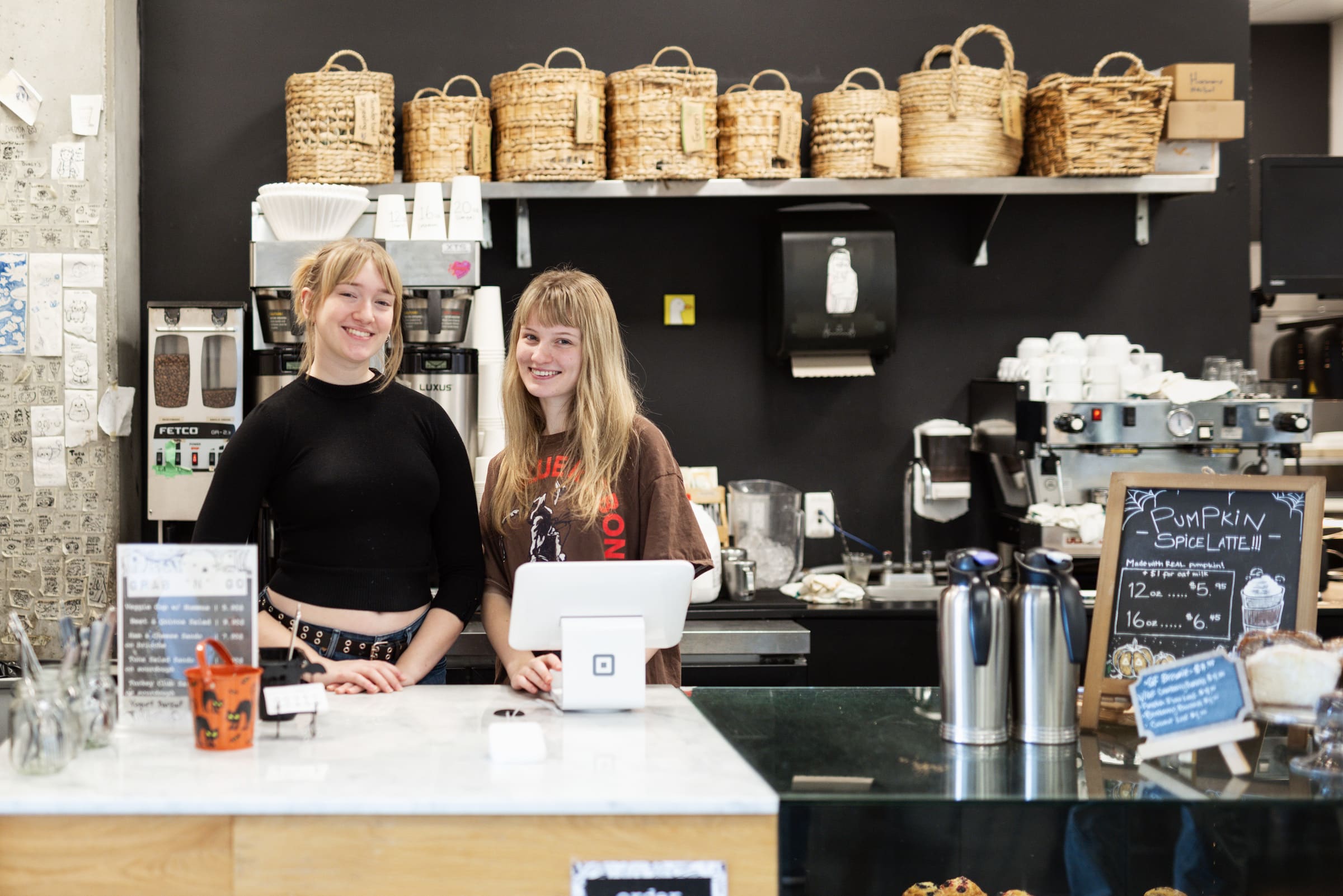 Two student baristas smile behind the counter of the campus café, with coffee equipment and woven baskets on shelves in the background.
