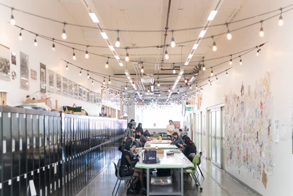 Students walk through an ECU hallway decorated with posters and artwork, lit by string lights overhead.