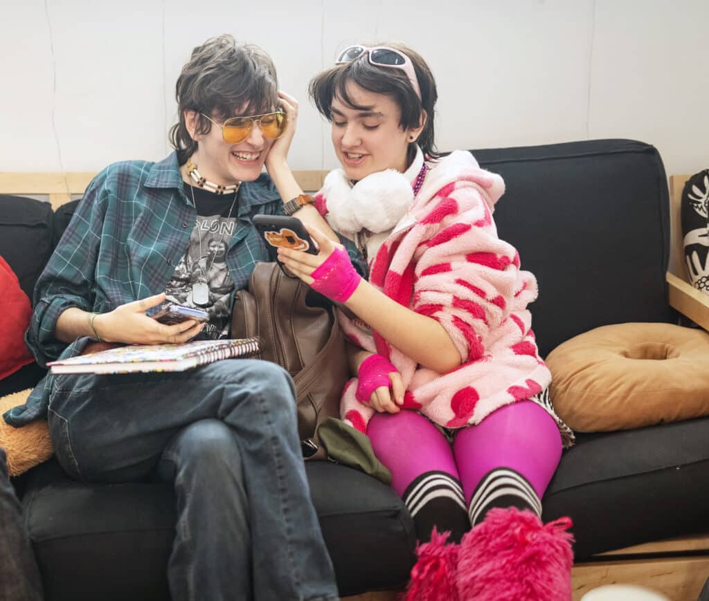 Two students in playful outfits sitting together on a couch, smiling while looking at a phone.