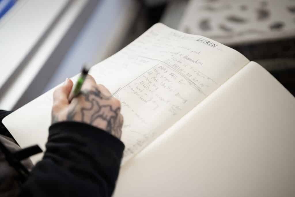 Close-up of a tattooed hand writing in a notebook with a green pen. The page is filled with handwritten notes, sketches, and headings, capturing an in-progress brainstorming or planning session.