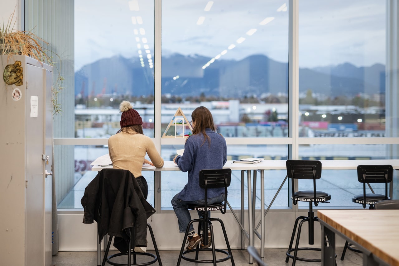 Two students working at a tall table with sketchbooks open, facing a large window with a view of Vancouver’s mountains.