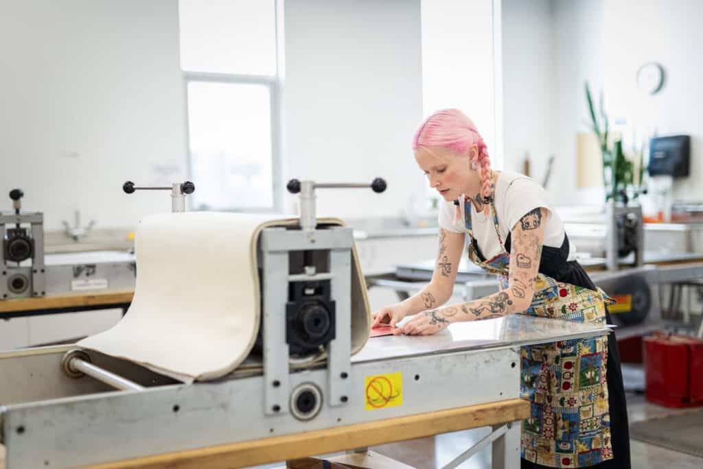 A student with pink hair and tattooed arms leans over a large printmaking press, carefully preparing a plate while wearing a patterned apron.