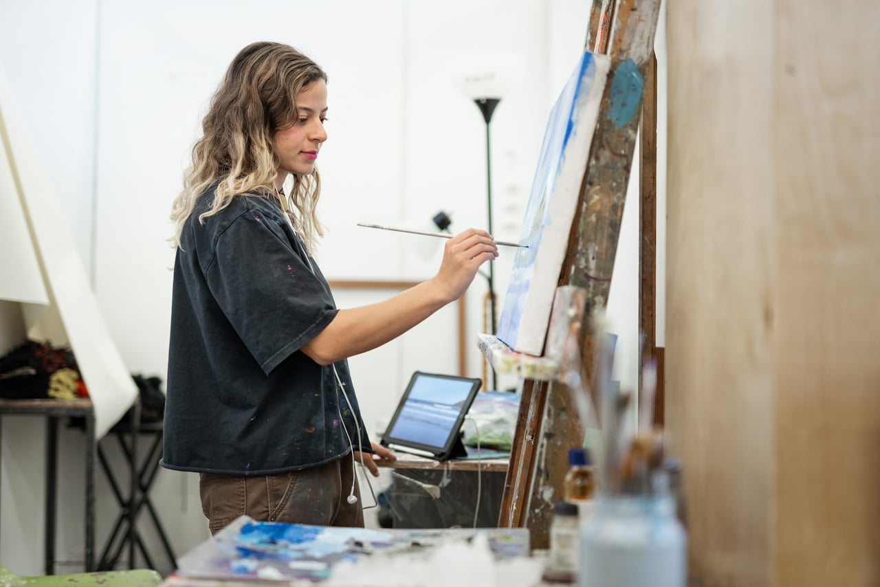 Student standing painting with a canvas propped up on an easel, using blue paint.