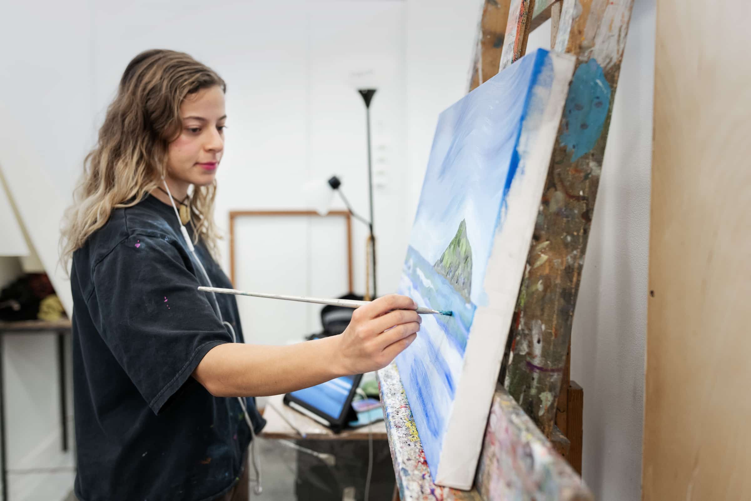 Student standing painting with a canvas propped up on an easel, using blue paint.