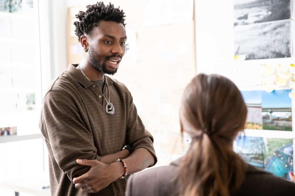 A man in a brown striped shirt with layered necklaces speaks to another person whose back is to the camera, in a bright room with photos and notes pinned to the wall.