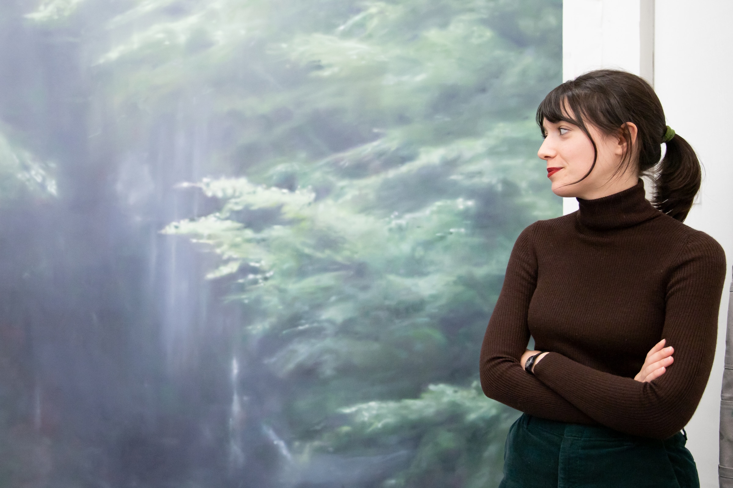 A woman with dark hair tied back, wearing a brown turtleneck sweater, stands with arms crossed, looking toward a large, blurred green-toned painting or mural in the background.