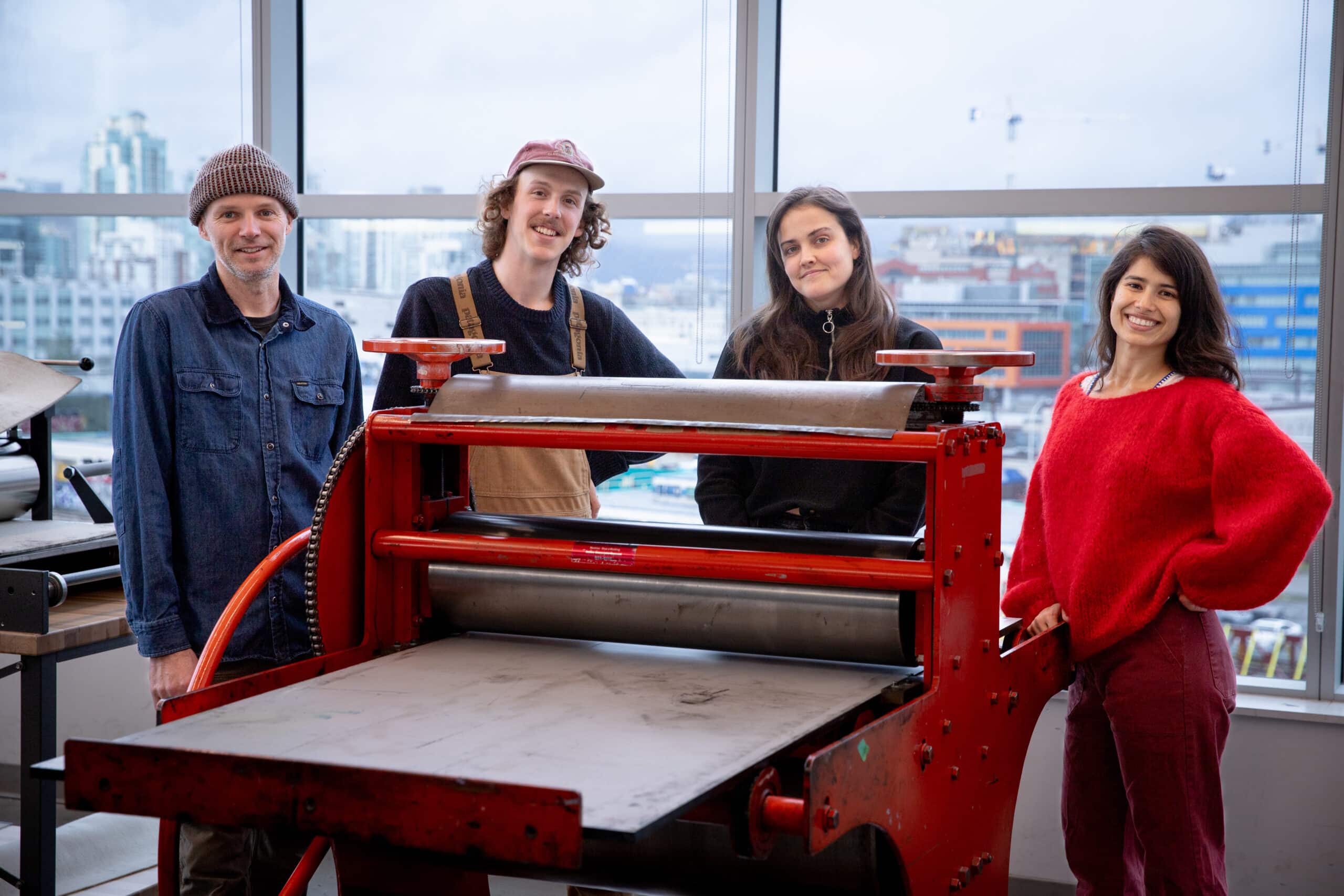 Group of four people standing behind a large red printmaking press in a bright studio with city buildings visible through the windows in the background.