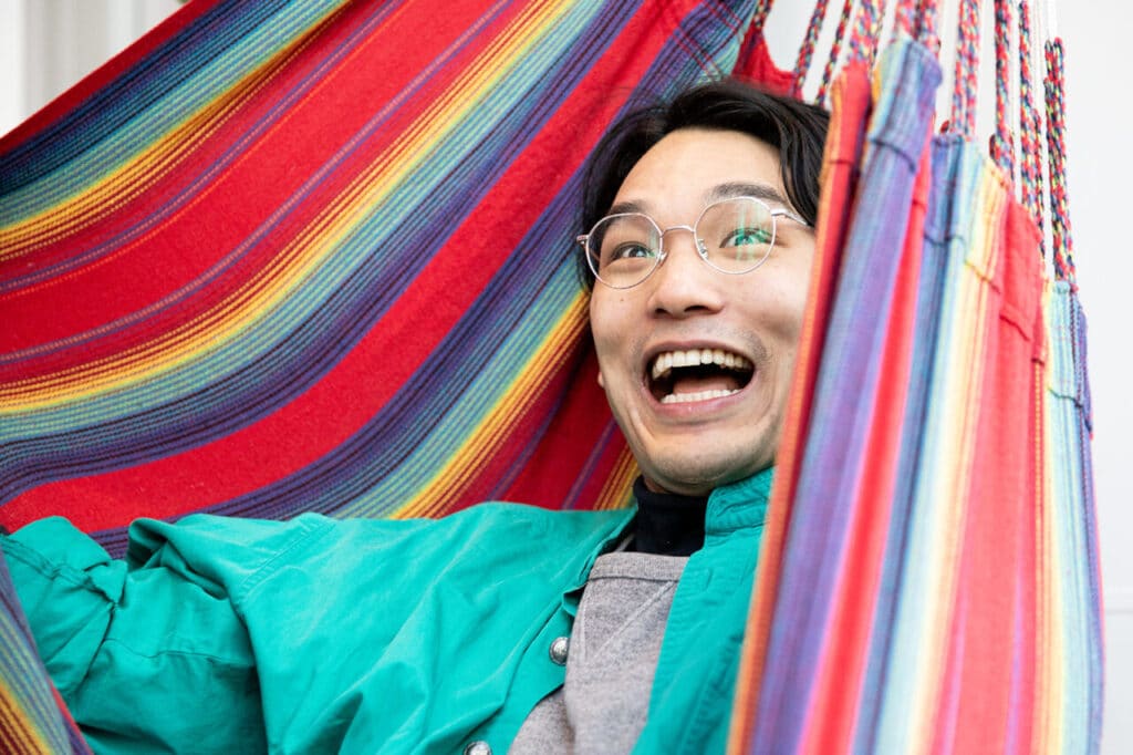 Person smiling widely while sitting in a colourful striped hammock, wearing glasses and a bright green jacket.