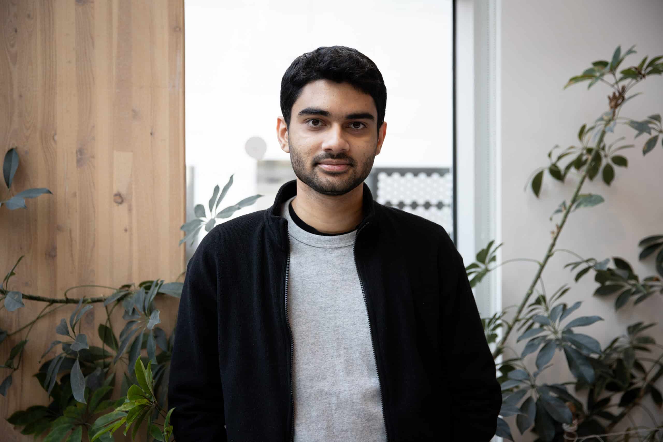 A person wearing a light grey shirt and black jacket stands indoors near a wooden wall and leafy plants, looking directly at the camera with a neutral expression.