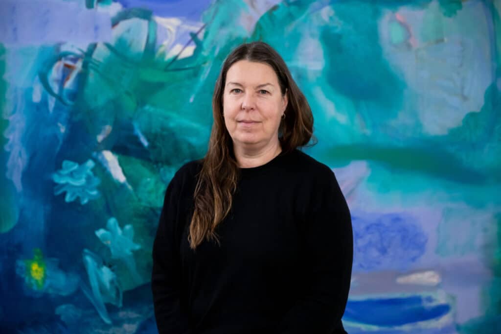 Portrait of a woman in a black sweater standing against a large abstract painting in blue and green tones.