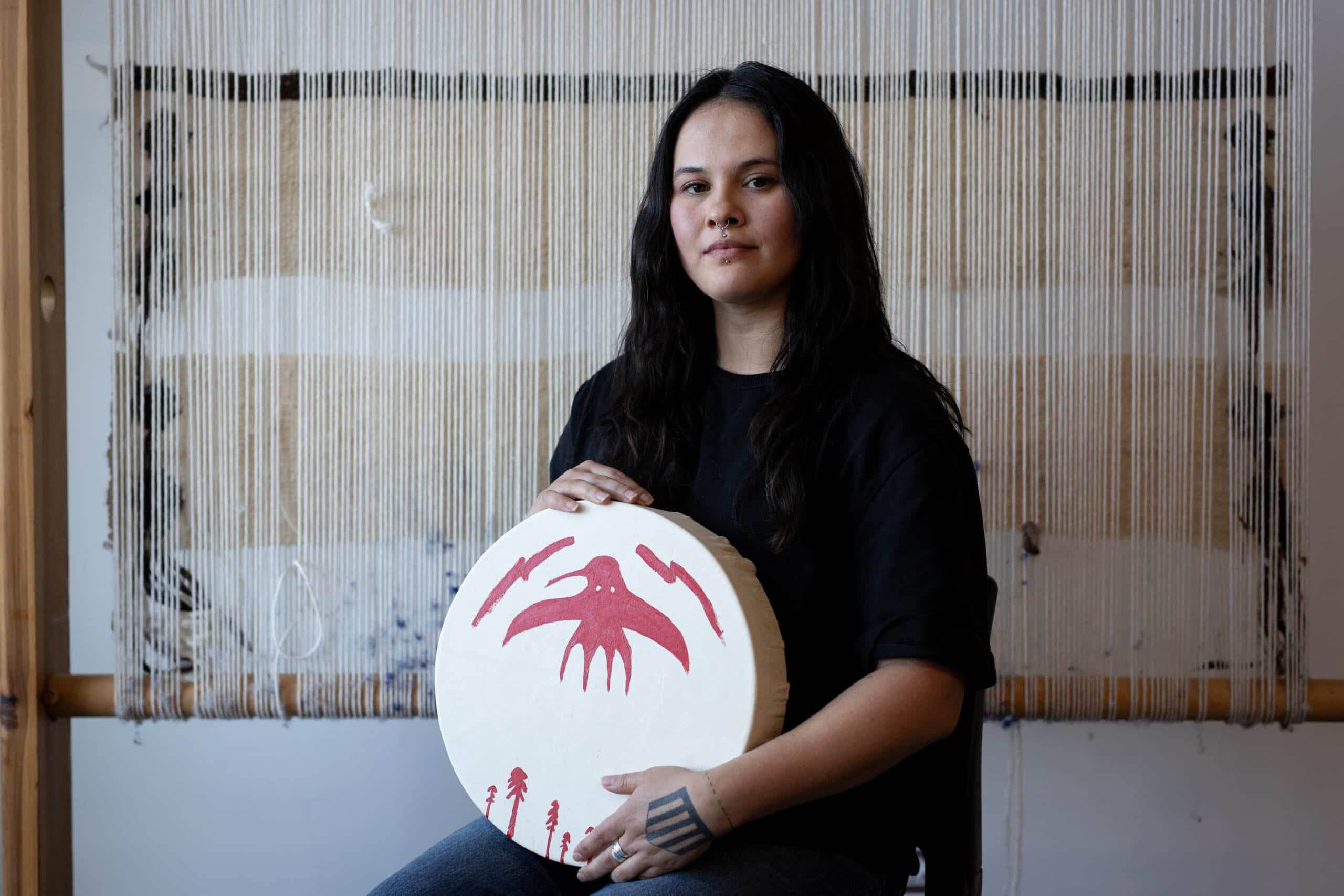 A person with long dark hair sits in front of a weaving loom, holding a white hand drum painted with a red bird and trees.