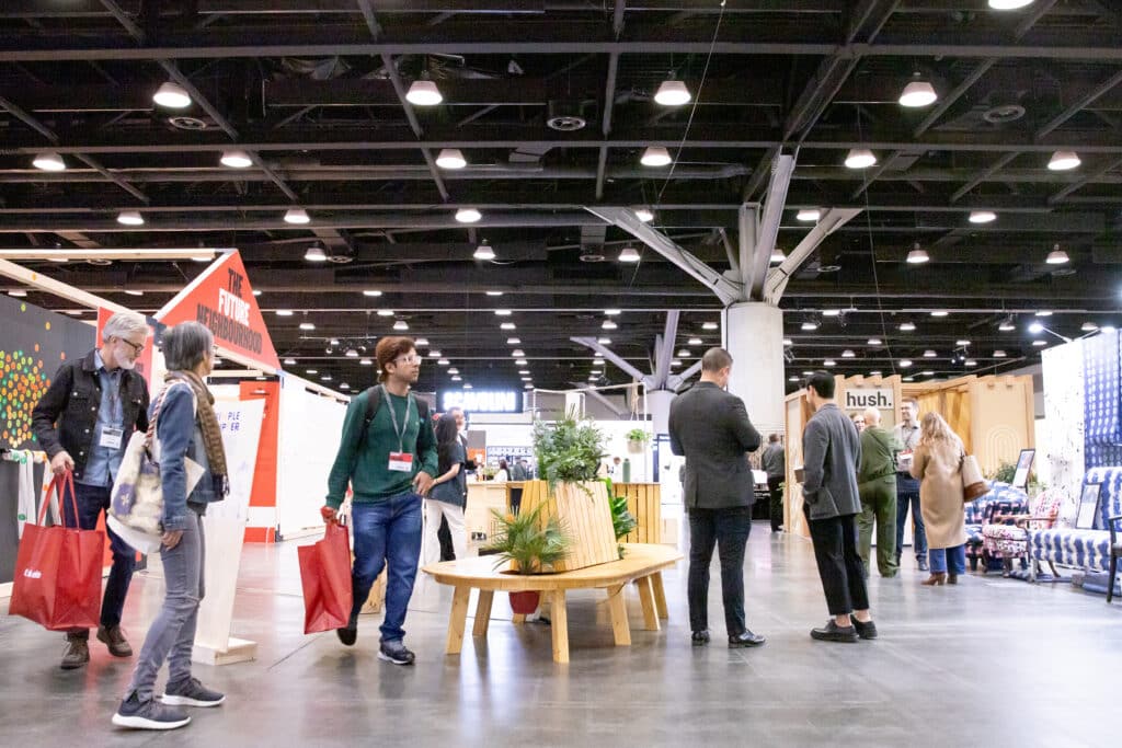Wide shot of trade show floor with people walking past booths, including The Future Neighbourhood installation and the hush. display.