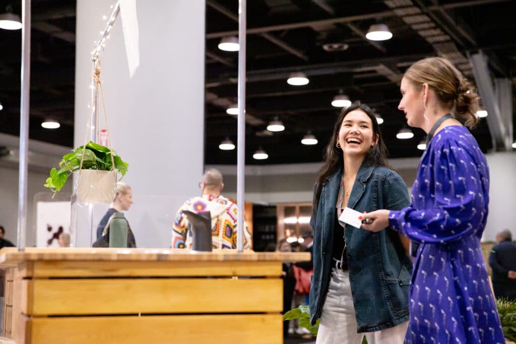 Two people smiling and talking beside a wooden installation with hanging plants.