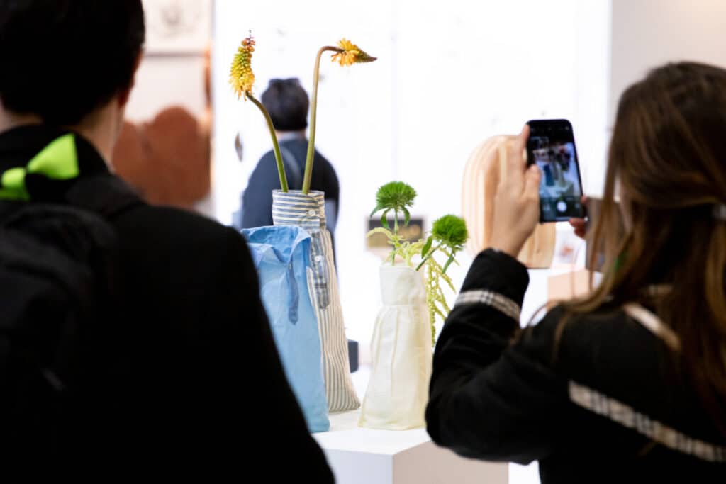 People viewing and photographing fabric-covered vases holding flowers at an exhibition.